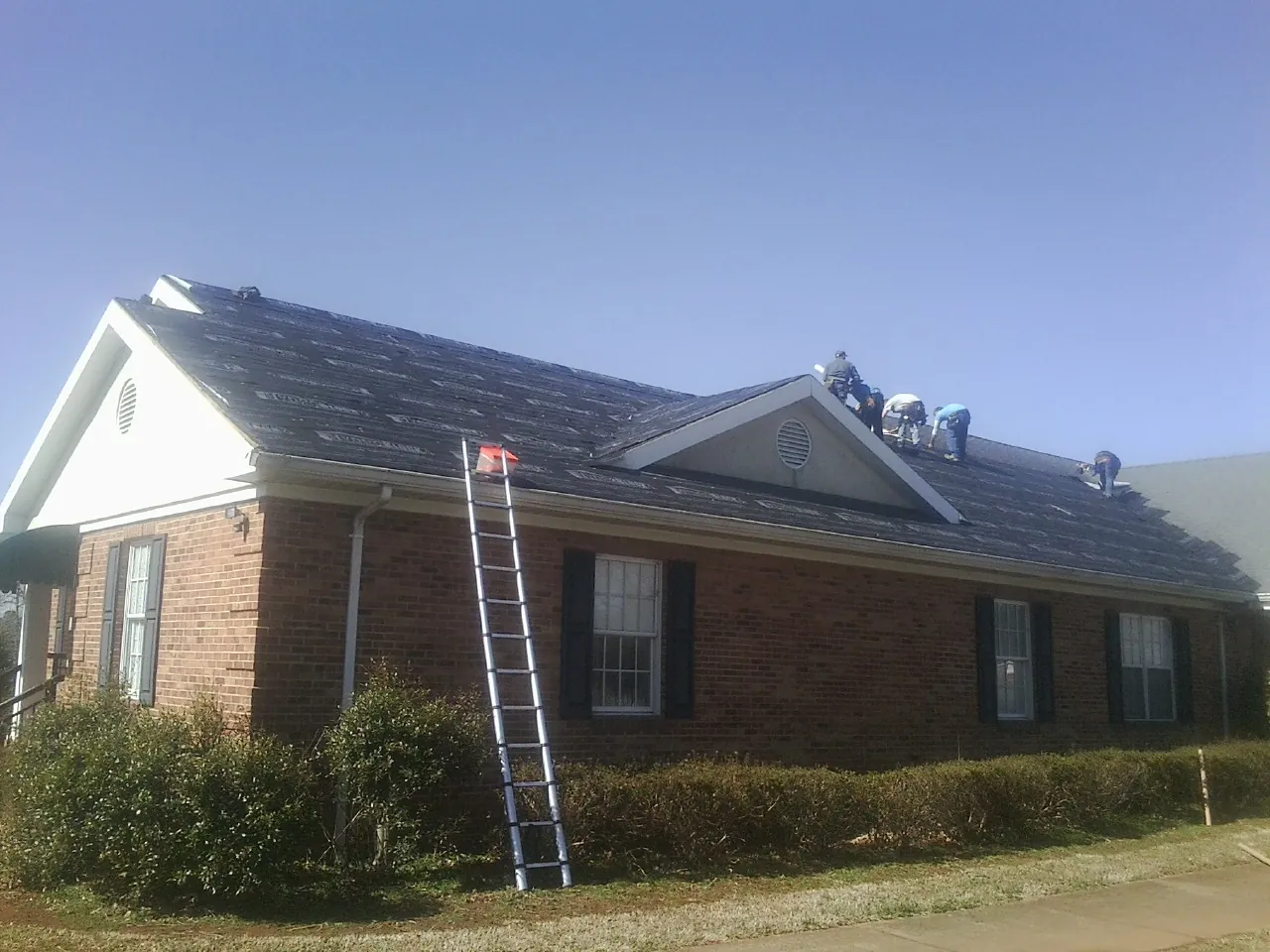 Crew tearing shingles off a brick ranch roof with a ladder set — Emergency Roof Repair work by our crew in Slickville