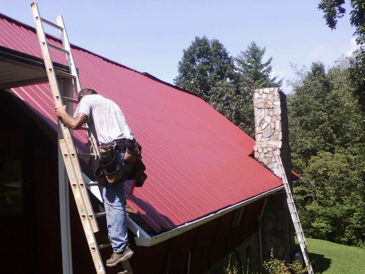 Roofer on a ladder beside a red metal roof and stone chimney — Soffit & Fascia Repair by Roofing Brothers in Slickville